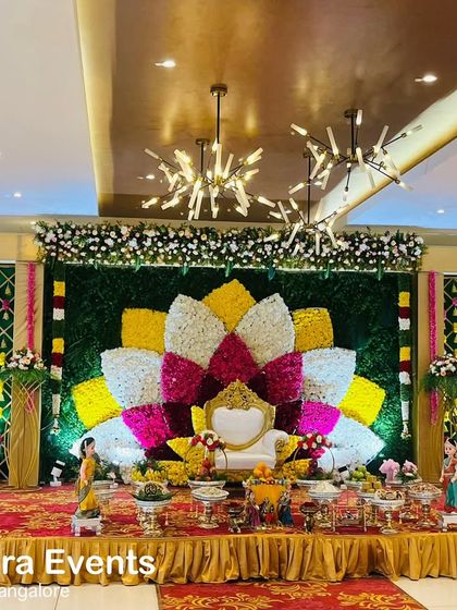 A wide-angle view of a magnificent Seemantham stage. The decor features a multi-colored floral lotus, decorative side panels, and a beautifully arranged display of traditional offerings.