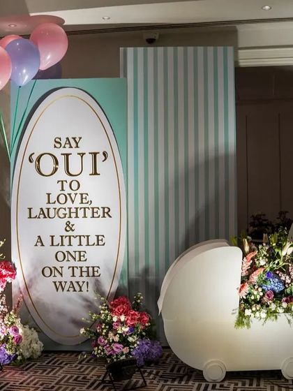 An elegant and unique baby shower backdrop with the message 'Say OUI to love, laughter & a little one on the way', complete with a pram prop filled with flowers.