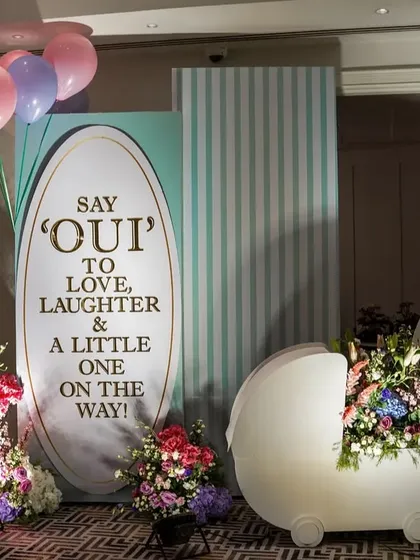 An elegant and unique baby shower backdrop with the message 'Say OUI to love, laughter & a little one on the way', complete with a pram prop filled with flowers.