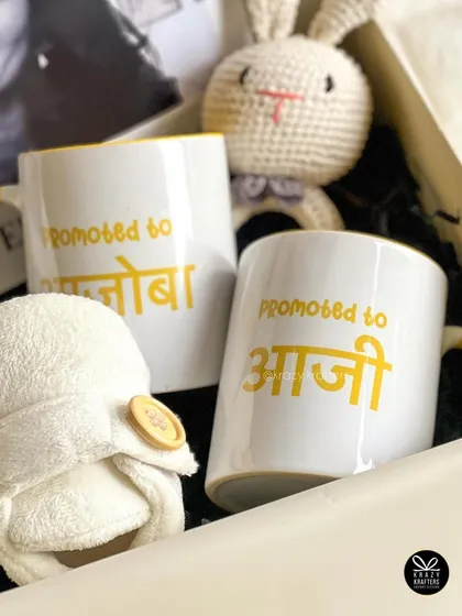 A close-up of the 'Promoted to Aaji & Ajoba' mugs, nestled next to a crochet bunny rattle and soft baby booties. It’s a box full of texture and warmth.