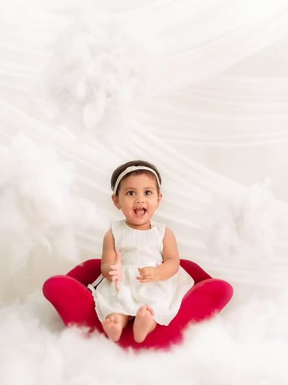 A toddler in a white dress sits on a red chair surrounded by fluffy white clouds, creating a "heavenly" themed portrait.
