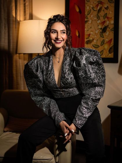 An indoor portrait of Ragini Dwivedi in a stylish silver and black top. The warm, soft lighting from the lamp creates an intimate and sophisticated mood.