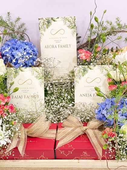 Here you can see how the invitation cards are nestled amongst the flowers and gifts. Each card can be personalized for different families, like the Arora, Bajaj, and Aneja families shown here, making every guest feel personally welcomed.