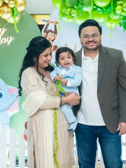 A happy family portrait at a jungle-themed first birthday party, with the parents and birthday boy posing together in front of a vibrant green and gold balloon backdrop.