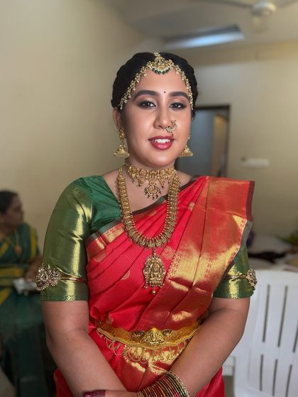 A full-length view of Dr. Indhu's bridal look. Her red and green Kanjeevaram saree, paired with heavy gold jewellery and flawless makeup, creates a stunning and regal appearance.