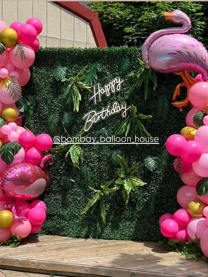 An outdoor flamingo-themed party with a green hedge wall backdrop, a neon sign, and bright pink balloon garlands.