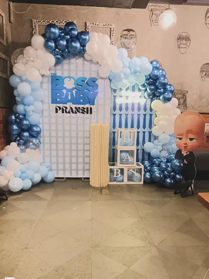 A fun "Boss Baby" themed decoration for Pransh. The setup includes a character cutout, a city-grid backdrop, and a blue and white balloon arch, perfect for celebrating a little boss.