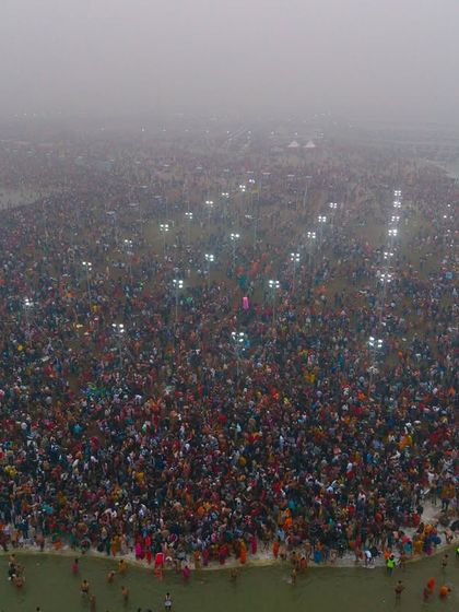 An aerial view of the Kumbh Mela grounds at dawn, with fog covering the river and lights illuminating the massive gathering of people.