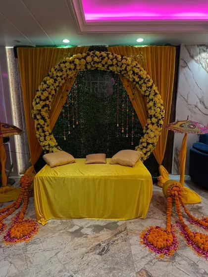 A circular floral arch made of yellow flowers creates a stunning focal point for this haldi ceremony. The setup includes a green backdrop, traditional props, and comfortable seating.