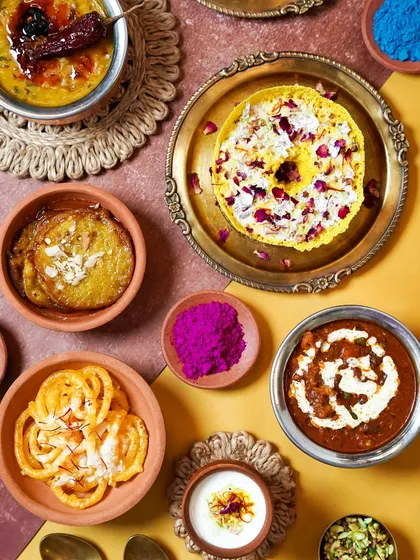 A vibrant spread of food for a Holi brunch, featuring both savory dishes and traditional sweets. The pops of color from the food and the scattered gulal capture the festival's joyful essence.