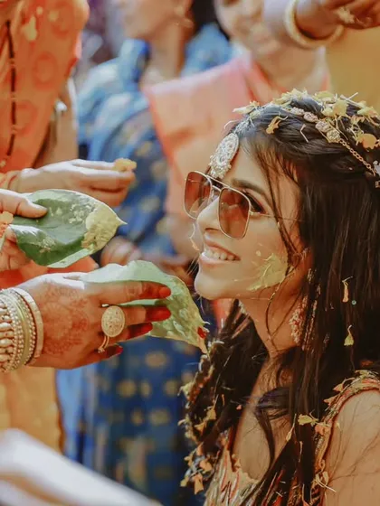 A cool bride enjoys her Haldi ceremony, wearing sunglasses as her family applies the turmeric paste.