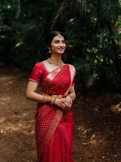A full-length shot of the bride in a beautiful outdoor setting. The red saree stands out beautifully, and her makeup is kept classic and timeless.