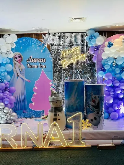 A magical Frozen theme for a first birthday party, featuring Elsa, a shimmer wall, and a winter wonderland of blue, purple, and silver balloons.