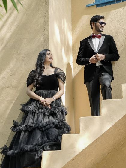 A stylish shot of a couple on a staircase, him in a tuxedo and her in a tiered black gown, creating a modern and chic pre-wedding look.