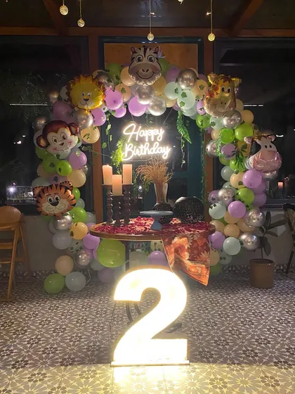 A jungle-themed 2nd birthday party at night. The circular balloon arch is decorated with animal foil balloons and a 'Happy Birthday' neon sign.