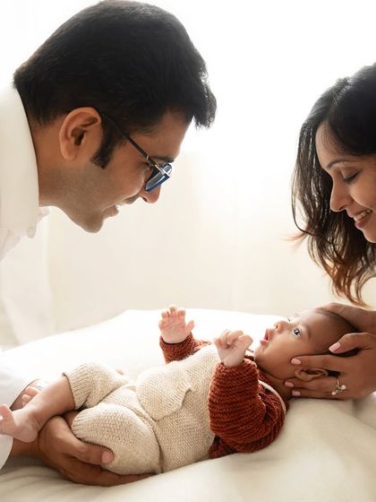 The first family portrait. Parents gazing lovingly at their new baby, who is dressed in a cozy knit outfit. A bright, light-filled image of pure happiness.