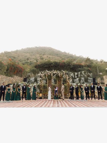 A wide shot of a beautiful outdoor vow ceremony, with the wedding party flanking the couple. Set against the Aravalli mountains, this image showcases the scale and beauty of a destination wedding ceremony.