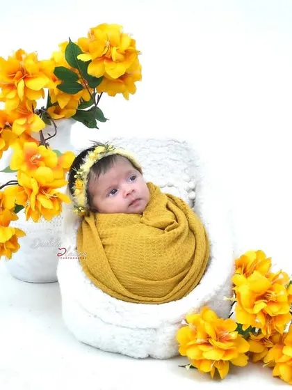 An adorable newborn, wrapped in a mustard yellow swaddle and matching headband, looks directly at the camera. The setup is surrounded by bright yellow flowers.