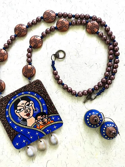 This piece, named 'Mamta', is a celebration of a mother's love. The hand-painted pendant shows a tender moment between mother and child, paired with matching wooden jhumkas and finished with delicate shells that add a soft, natural touch.