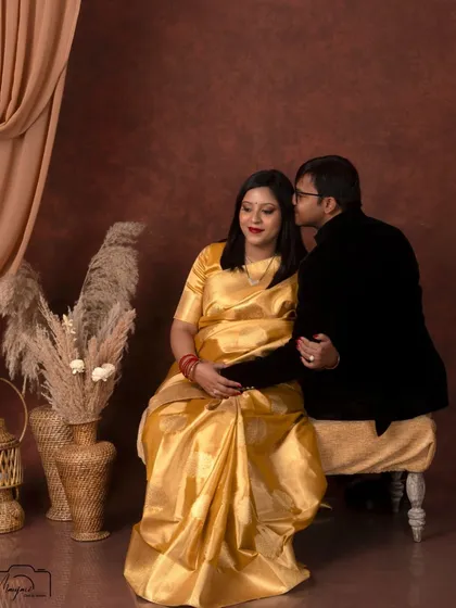 A tender moment between this couple, seated in a classic studio setup. The golden saree and his black velvet jacket create a look of pure elegance.
