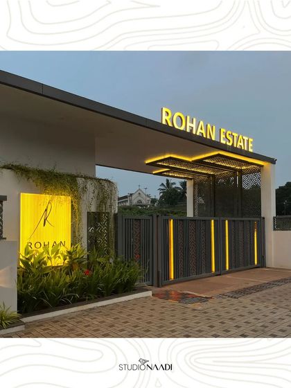 The illuminated entry gate for Rohan Estate in Mangaluru, seen at dusk. The design combines modern perforated panels, warm backlighting, and integrated vertical light strips on the gate to create an elegant and secure entrance that is visually appealing day and night.