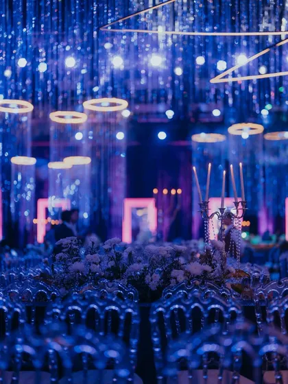 A beautifully blurred shot of the sangeet decor, focusing on the shimmering lights and crystal elements. The ghost chairs and floating candle centerpieces are bathed in blue light, creating a dreamy and ethereal atmosphere for the celebration.
