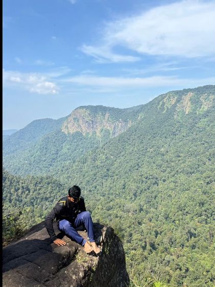 A trekker sitting on a cliff edge, looking out at the vast green valley of Agumbe.