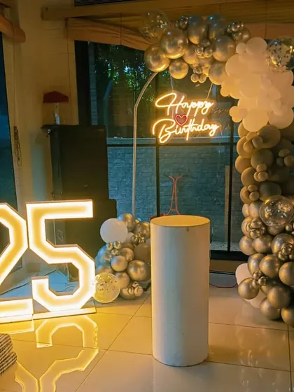 A 25th birthday party with a silver and white balloon arch, a 'Happy Birthday' neon sign, and a large '25' marquee light.
