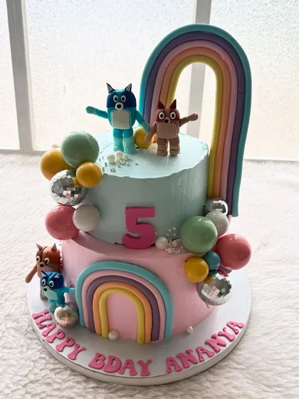 A colorful two-tier Bluey-themed cake for a 5th birthday party. It features handcrafted characters, rainbows, and fun disco balls to bring the cheerful world of Bluey and Bingo to life.