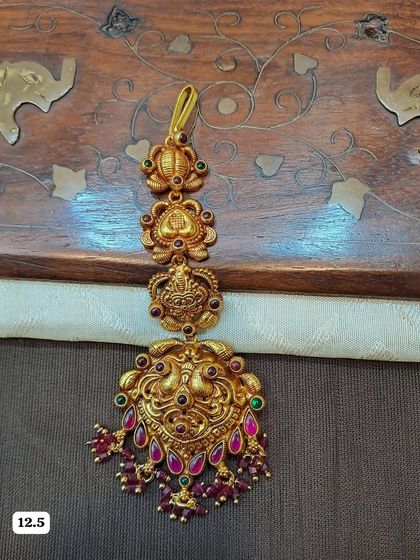 This traditional maang tikka features a detailed peacock design and is crafted from 92.5 silver with gold plating. The ruby-colored bead drops make it perfect for bridal wear.