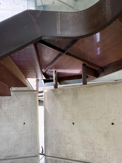 The underside of a perforated steel staircase in the 'House of Stories'. The detail shows the honest expression of materials, with the structure of the stair left exposed, creating intricate patterns of light and shadow on the concrete walls below.
