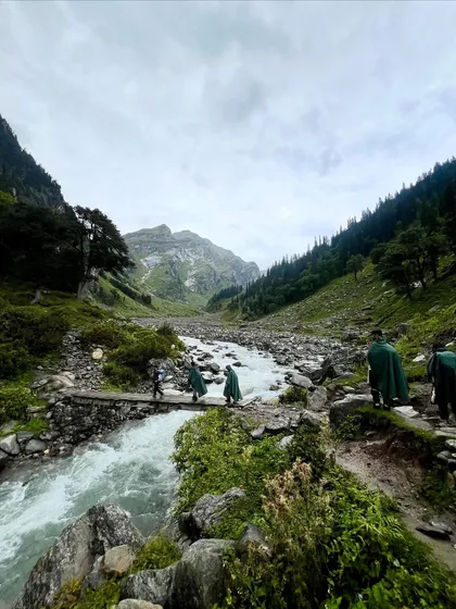 Trekkers in ponchos crossing a river on a rustic wooden bridge. Rain or shine, the adventure on the Hampta Pass never stops.