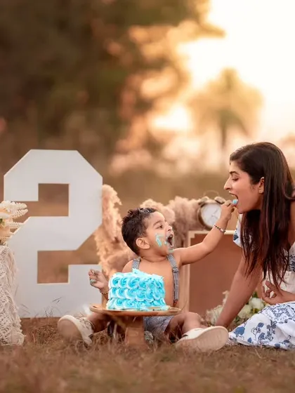 A fun-filled outdoor cake smash session for a second birthday, showing a mother feeding her son a piece of blue cake in a beautiful park setting.