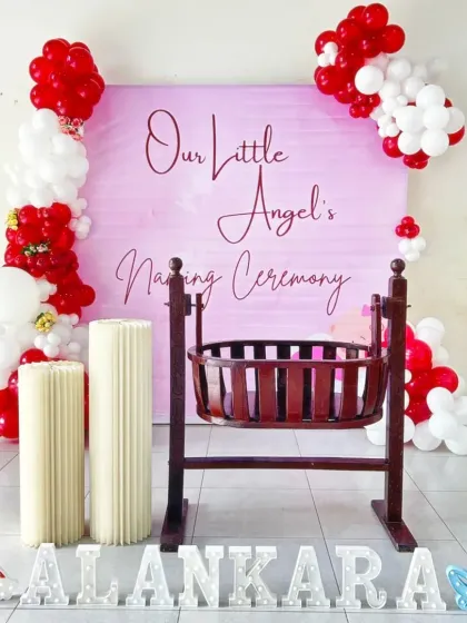 A sweet and simple naming ceremony for a baby girl. The pink backdrop is framed by red and white balloons, with a traditional wooden cradle as the centerpiece and marquee letters spelling the name.