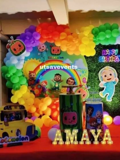 A vibrant Cocomelon-themed birthday with a rainbow-colored balloon arch and a grass wall backdrop. The setup includes a Cocomelon school bus prop and light-up letters for the name 'AMAYA'.