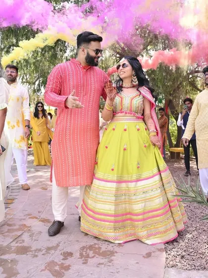 A grand entrance for the Haldi ceremony. The couple walks in surrounded by friends and family holding colorful smoke bombs, creating a vibrant and energetic start to the festivities.