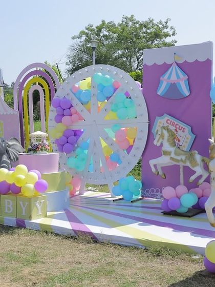 A wide view of the outdoor carnival stage, showcasing the full setup with a Ferris wheel, carousel horses, and pastel balloon decorations under the bright sun.