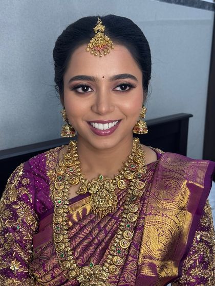 A full-face portrait of Anitha, showing the complete, balanced makeup look. Her features are beautifully enhanced, and the entire look comes together with her stunning gold jewellery and embroidered blouse.