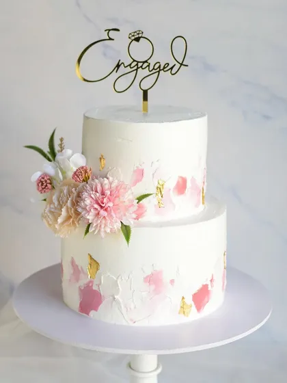 This two-tiered engagement cake features a beautiful textured buttercream effect with strokes of pink and gold leaf. The design is finished with fresh flowers and a custom "Engaged" topper.