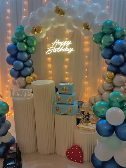 A clear view of the birthday balloon arch with the "Happy Birthday" neon sign glowing brightly. The plinth stands are ready for the cake and gifts.