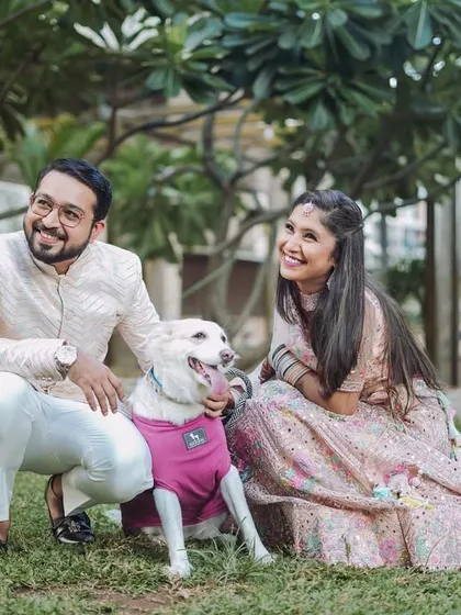 A happy couple in elegant ethnic wear poses with their adorable white dog in a park. This photo shows that I love including your furry family members in your couple session to make the memories even more special.