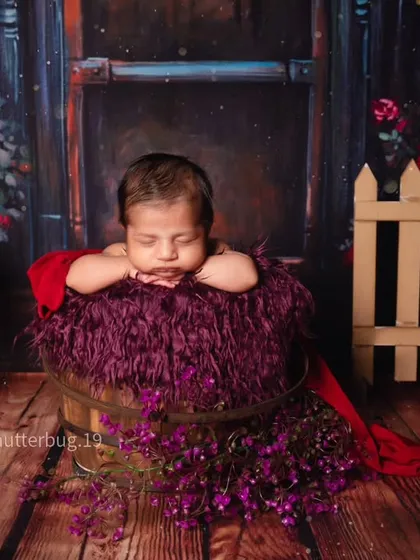 A moody and romantic floral theme. The baby rests in a bucket filled with rich purple fur, set against a dark wood floor and a backdrop of deep red roses.