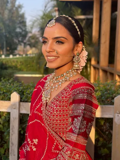 A breathtaking bride captured in the soft, natural light of her outdoor wedding. The makeup is designed to be luminous and fresh, complementing her red lehenga and floral bun.