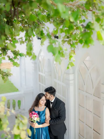 A beautiful portrait of the groom kissing the bride's forehead, framed by lush green leaves against a white architectural backdrop.