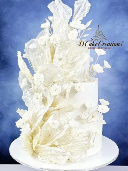 A modern and artistic cake for a 10th-anniversary celebration. This three-tiered white cake is adorned with abstract, delicate rice paper sails and scattered white sugar flowers for a contemporary, ethereal look.
