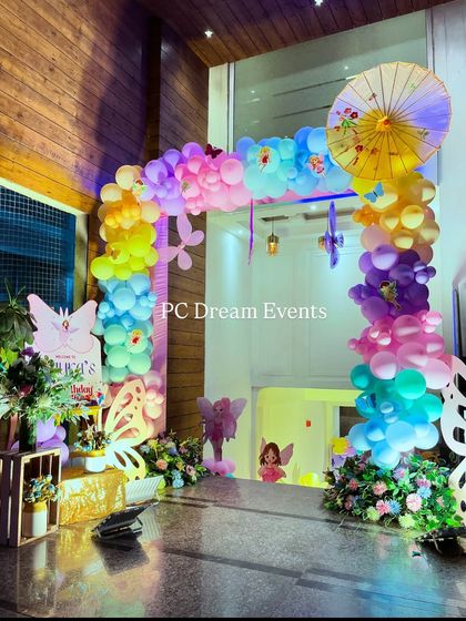 A whimsical butterfly and fairy-themed birthday entrance. A rainbow balloon arch, accented with a Japanese umbrella and butterfly cutouts, creates an enchanting welcome for Aarya's first birthday.