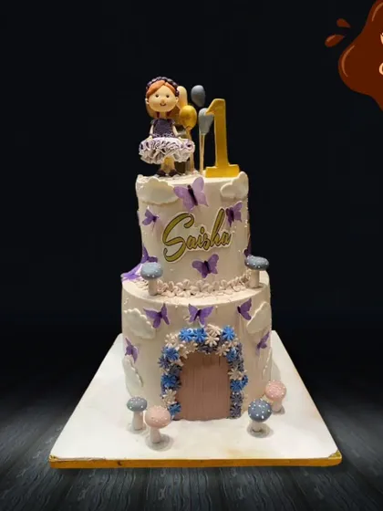 A beautiful two-tier cake for a first birthday, decorated with delicate purple butterflies and a floral archway. A sweet figurine of a little girl sits on top, making it a whimsical and personal creation.