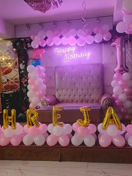 A first birthday stage setup in pink and white, with a large throne-like chair for the birthday girl and her name in foil letters.
