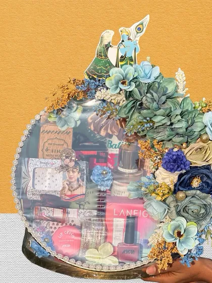 A modern take on a Teej hamper, this round box is filled with cosmetics from brands like Laneige. The blue floral arrangement and Radha Krishna cutout on top add a traditional touch to a contemporary gift.