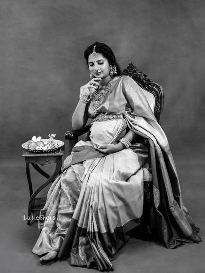 A contemplative black and white portrait of a mother-to-be in a traditional saree. Seated on an ornate chair, she looks down with a gentle expression, creating a classic and artistic feel.